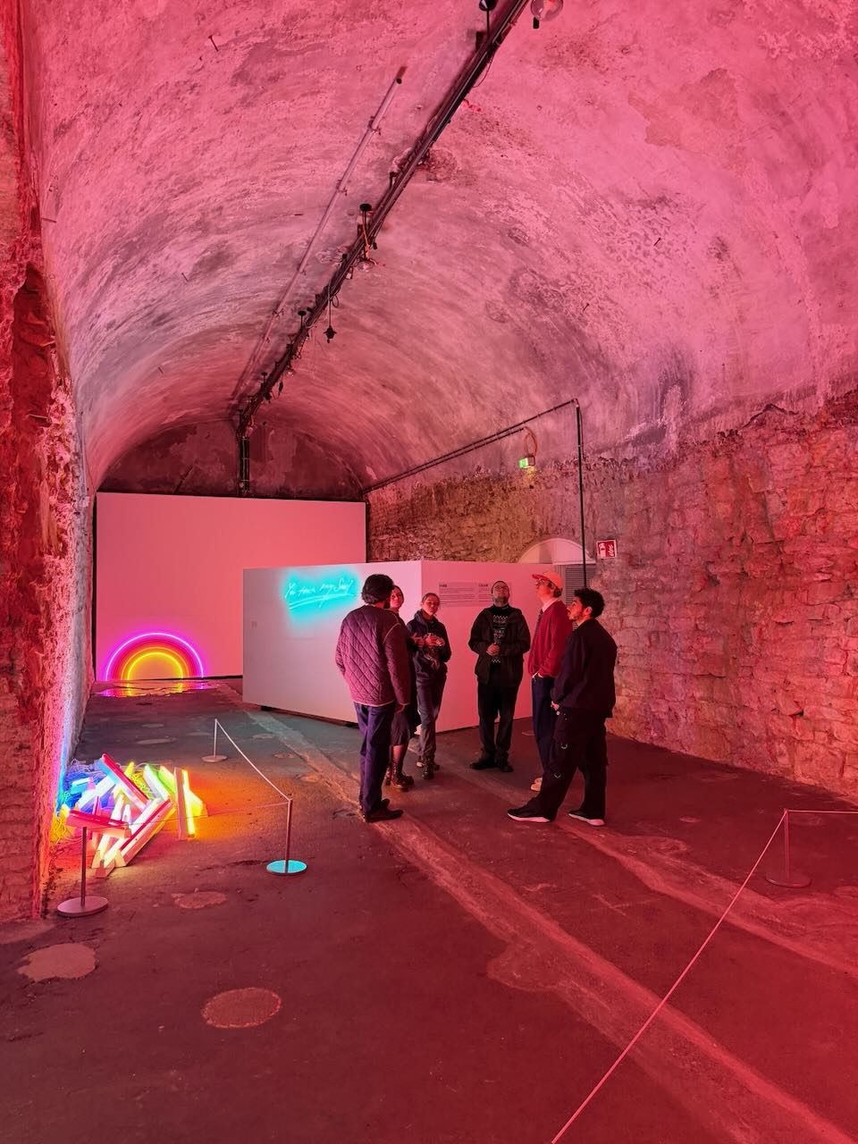 A group of students and staff on a guided tour in a vaulted brick cellar lit in pink and red, with neon works on the floor and on a white wall.