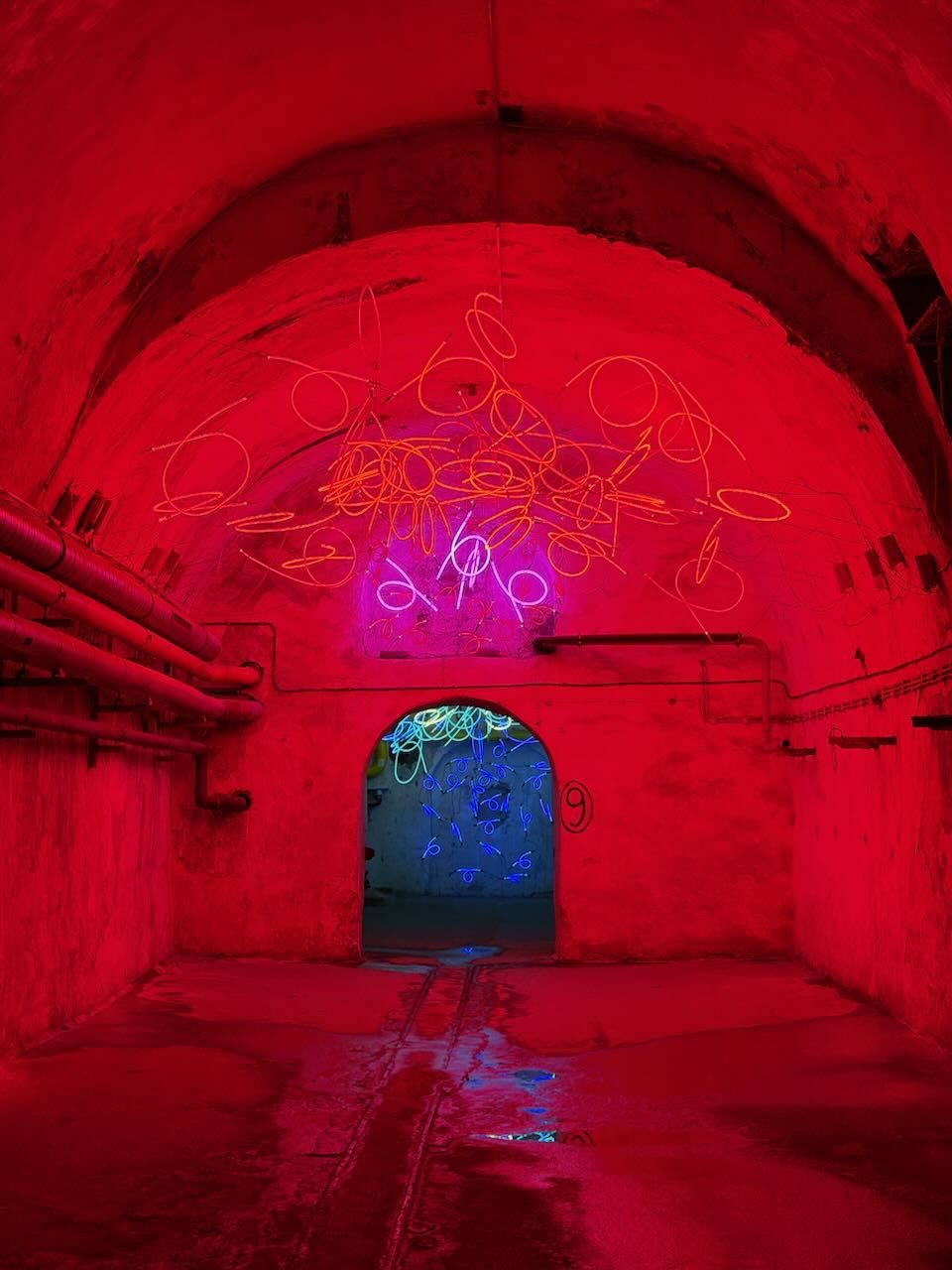 A cellar corridor bathed in red light, with loops of orange and pink neon tubing hanging from the vaulted ceiling and a doorway glowing blue at the end.