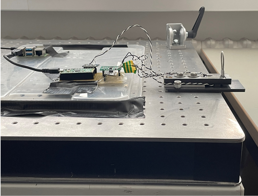 Laboratory bench with the test setup: a single-board computer and sensor boards wired to a load cell clamped to a perforated steel table, used to calibrate the weight measurement.